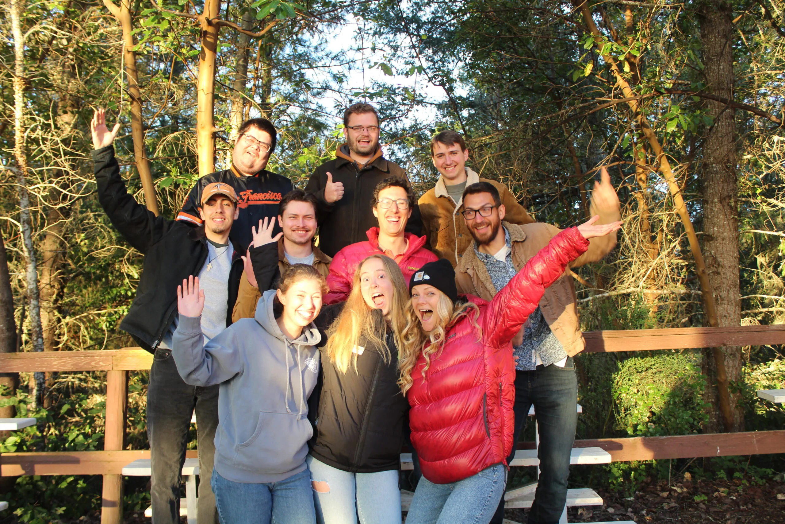 Ten people pose together outdoors on wooden benches, smiling and waving, surrounded by trees in bright sunlight.