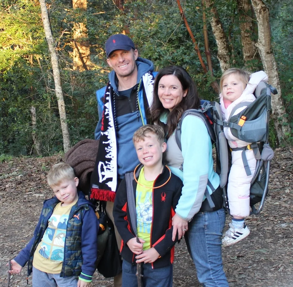 A family of five poses in a forest. Two adults stand with three children, one child is carried in a backpack carrier. They all appear happy and ready for a hike, surrounded by trees and sunlight filtering through.