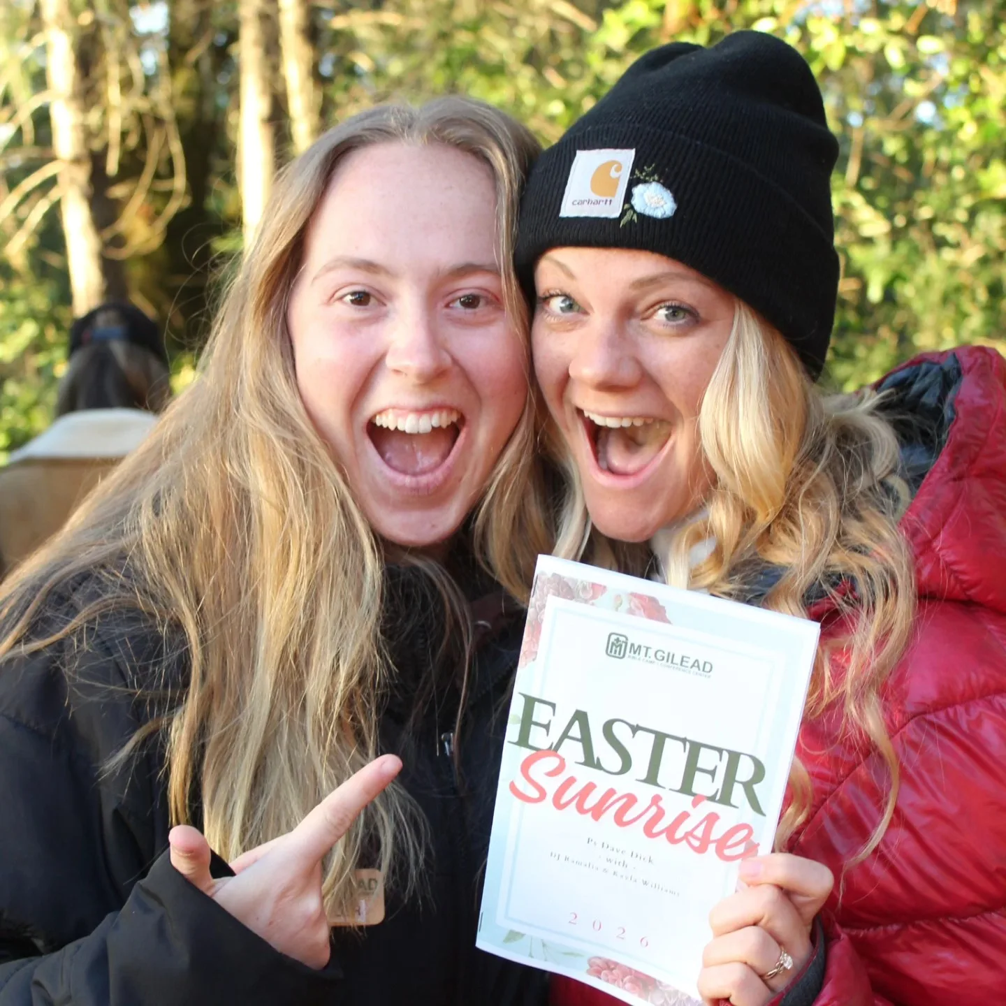 Two smiling women stand outdoors, one pointing to a program that reads Easter Sunrise with Mt. Gilead at the top. Both are wearing jackets, and one has a black Carhartt beanie. Trees are visible in the background.