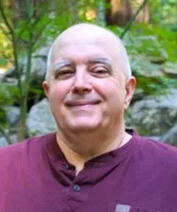 A bald man wearing a maroon shirt smiles outdoors, with greenery and rocks in the blurred background.
