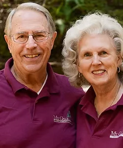 An elderly man and woman smile at the camera, both wearing matching maroon polo shirts, standing close together outdoors with greenery in the background.