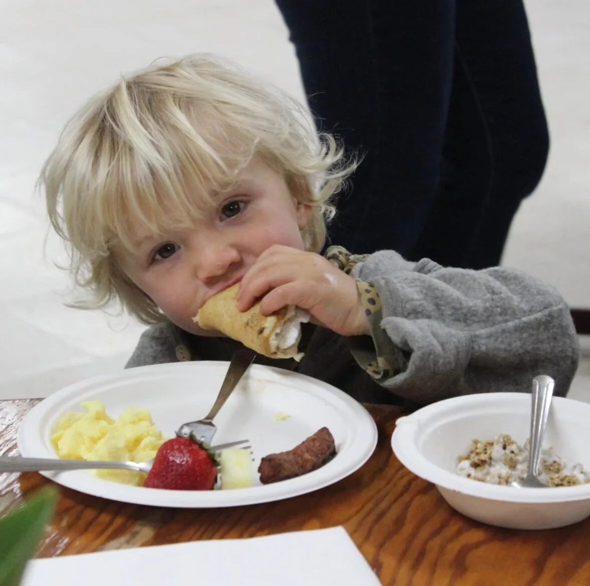 Young child with blonde hair eating a rolled pancake at a table, with a plate of scrambled eggs, strawberry, sausage, and a bowl of cereal in front of them.