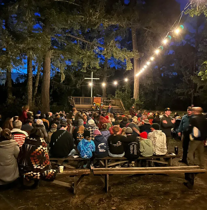 A large group of people sits on benches outdoors at night, facing a stage with a lit cross, under string lights and tall trees.