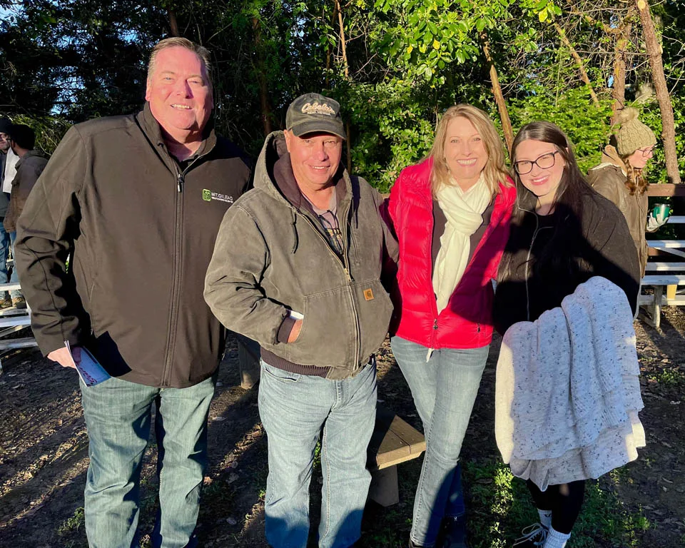Four adults stand outdoors in sunlight, smiling at the camera. They wear jackets and casual clothes. Trees and benches are in the background.