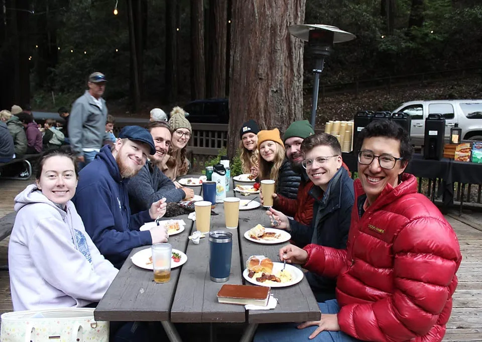 A group of people smile and enjoy a meal together at an outdoor wooden table surrounded by tall trees, wearing warm jackets and hats.