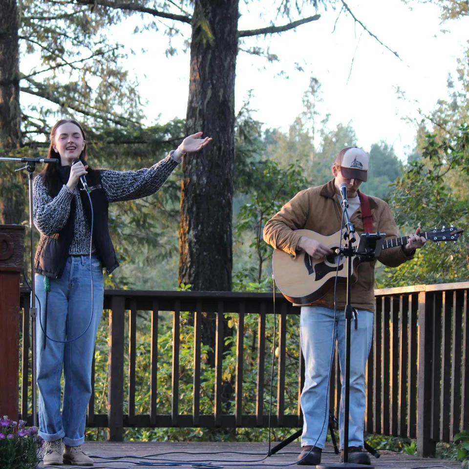 Two people perform on an outdoor wooden stage; one sings into a microphone with an arm raised, while the other plays an acoustic guitar. They are surrounded by trees and greenery under soft daylight.