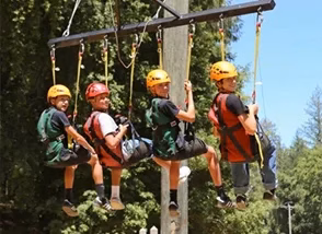 GuestRetreats-feature-goliath Group of children wearing helmets and harnesses sit on a suspended rope obstacle in a forest adventure course at a summer camp.