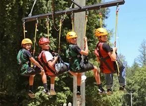 Group of children wearing helmets and harnesses sit on a suspended rope obstacle in a forest adventure course at a summer camp.