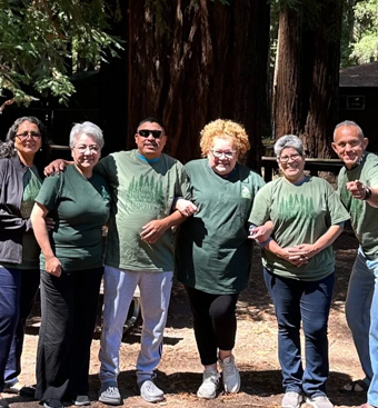 Group of six adults in green shirts posing outdoors, smiling with arms linked.”](https://)