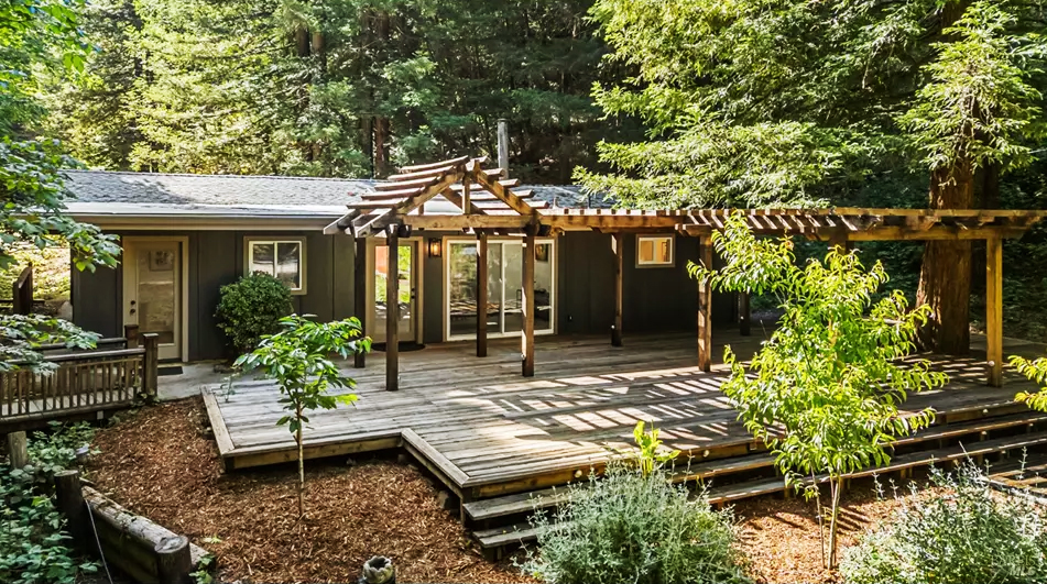 Wooden cabin with a large deck and pergola, set among tall trees in a forested yard.