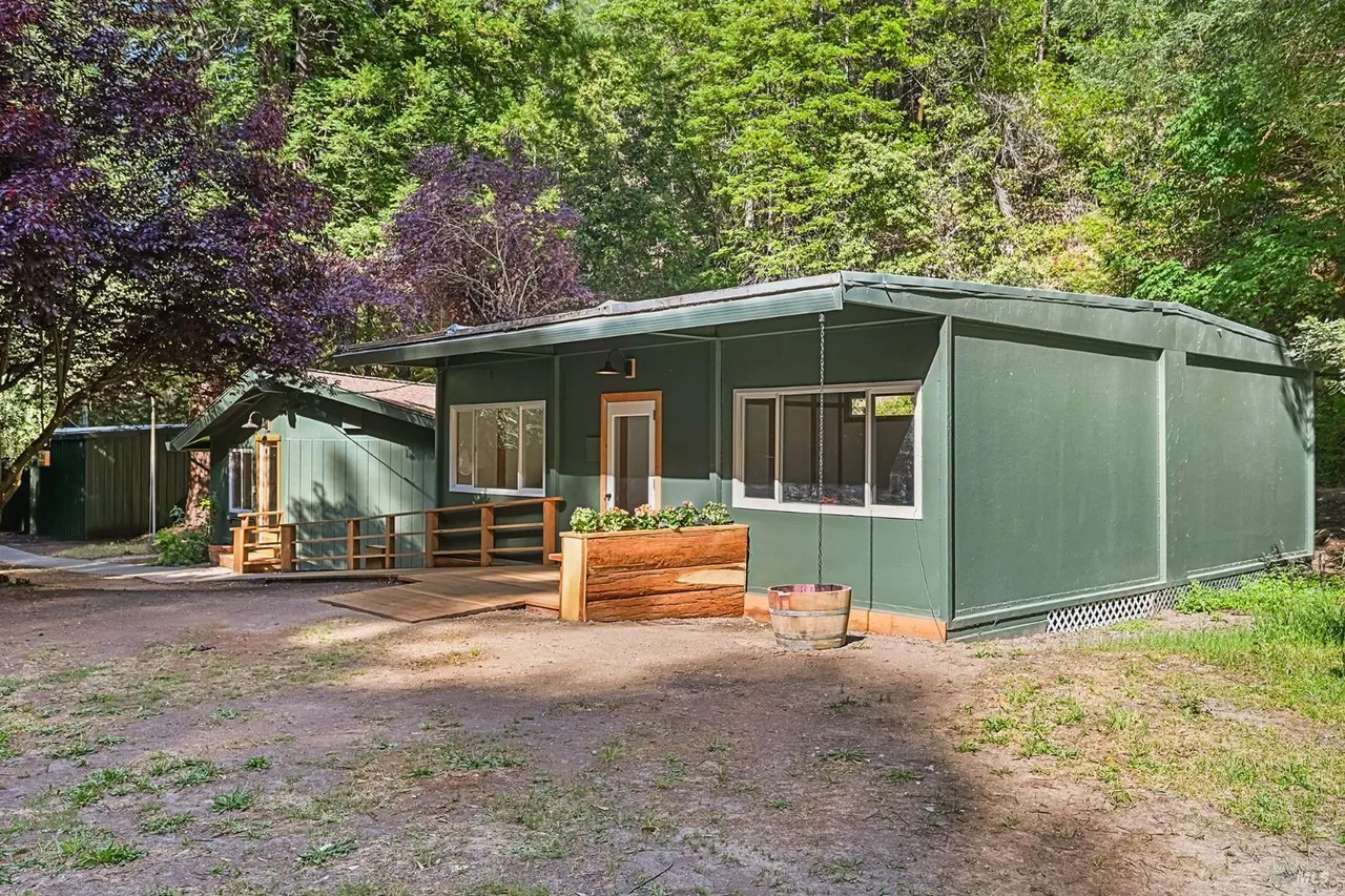 Green single-story cabin in a wooded clearing with a wooden porch and planters outside, bathed in sunlight.