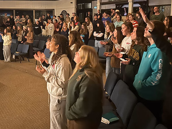 Large crowd of teenagers and young adults standing and clapping in a hall during an event. In the foreground, a few people face forward, cheering as others in the audience stand.