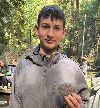 Young man outdoors in a forested area, wearing a gray zip-up jacket and holding a small round patch on his chest with his right hand.