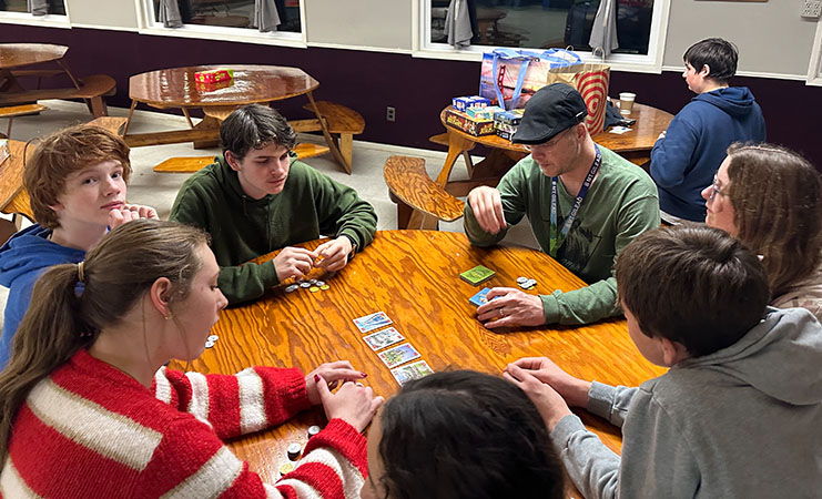 A group of eight teenagers sits around a wooden table indoors, playing a card or board game with cards and small tokens spread across the table.