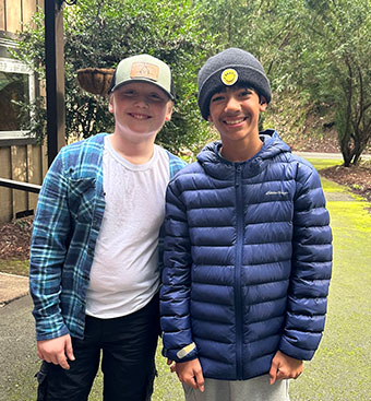 Two smiling teens standing outdoors on a tree-lined path; one wears a plaid shirt over a white tee with a cap, the other a blue puffer jacket and a beanie.