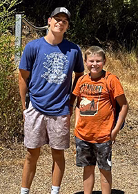 Two boys stand outdoors on a sunny day; one in a blue shirt and light shorts with a cap, the other in an orange shirt and dark shorts.