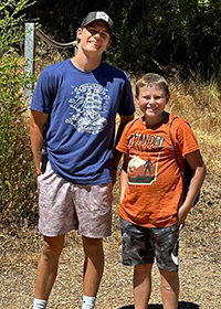 Two boys stand outdoors on a sunny day; one in a blue shirt and light shorts with a cap, the other in an orange shirt and dark shorts.