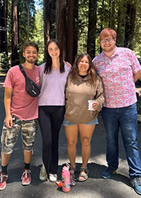 Four friends posing together in a forest, smiling at the camera; a pink water bottle on the ground nearby.