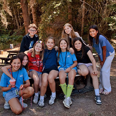 Group of smiling children at a camp outdoors, posing around a wooden table in a forest setting.