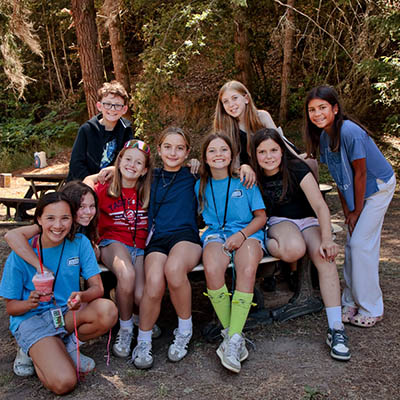 Group of smiling children at a camp outdoors, posing around a wooden table in a forest setting.