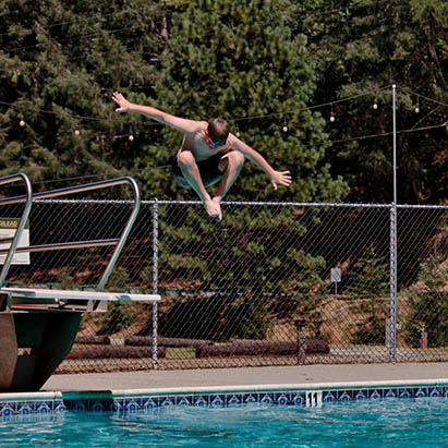 Young man mid-air diving from a pool diving board into a blue outdoor pool, with a chain-link fence and trees in the background.