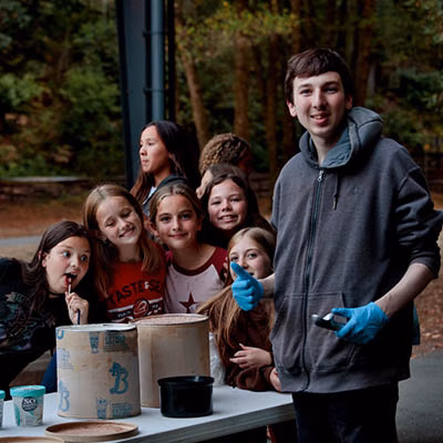 Group of kids gathered around a table outdoors, watching a young man in a gray hoodie with blue gloves lead an activity.