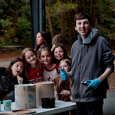 Group of kids gathered around a table outdoors, watching a young man in a gray hoodie with blue gloves lead an activity.