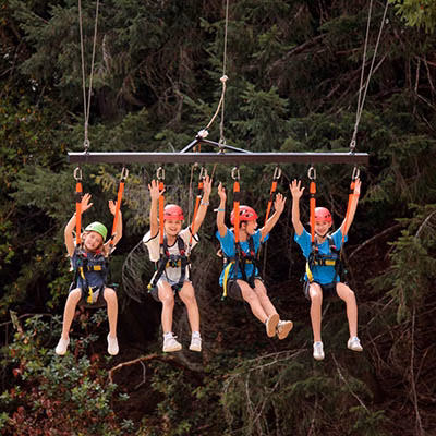 Four kids wearing helmets and harnesses zip-lining together through a forest, their arms raised in excitement.