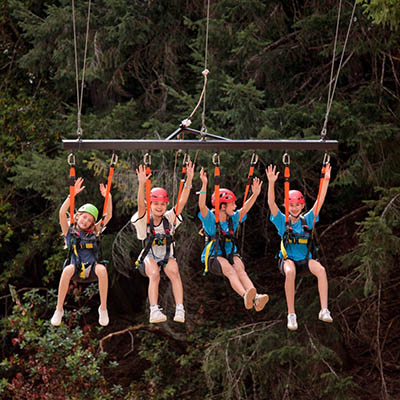 Four kids wearing helmets and harnesses zip-lining together through a forest, their arms raised in excitement.
