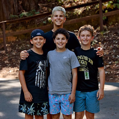 Four smiling boys posing outdoors with an adult behind them; a wooden railing and trees in a park setting.