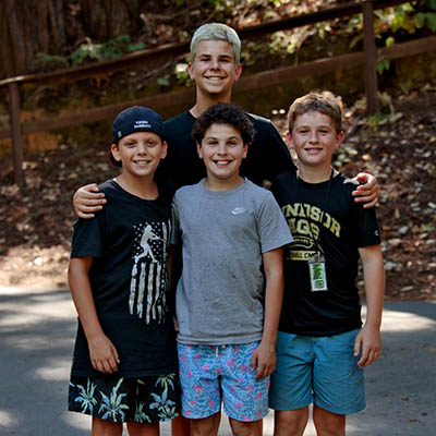 Four smiling boys posing outdoors with an adult behind them; a wooden railing and trees in a park setting.