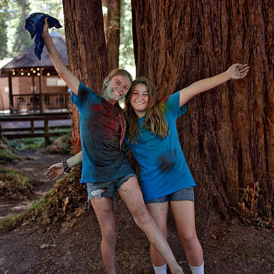Two smiling girls in blue shirts, muddy from a color run, lean against a large tree with their arms around each other outdoors.