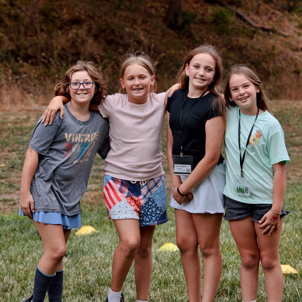 Four girls outdoors with arms around each other, smiling at the camera on a grassy field.