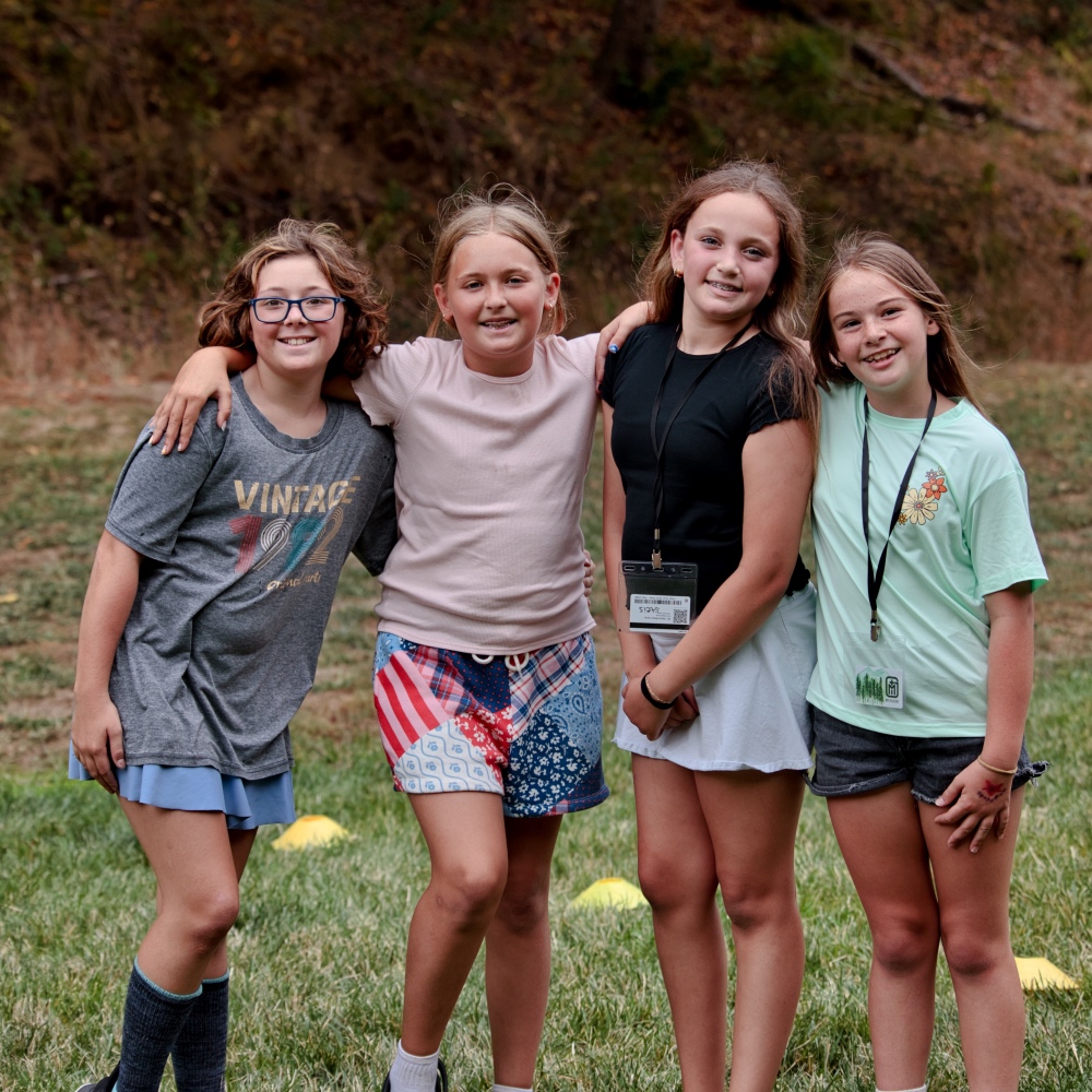 Four girls outdoors with arms around each other, smiling at the camera on a grassy field.