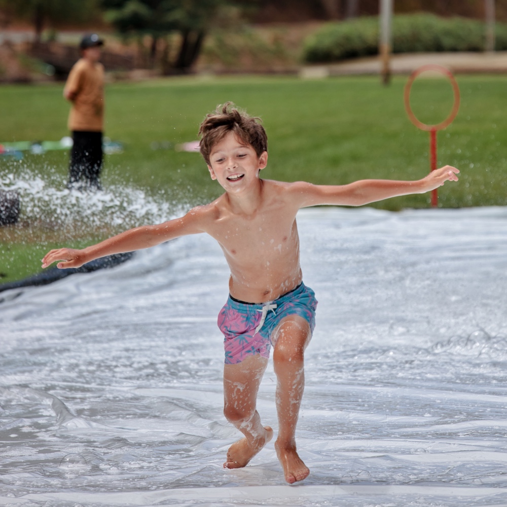 Young boy in colorful swim trunks runs on a wet slip-and-slide, water spraying around him in a park grassy field behind.