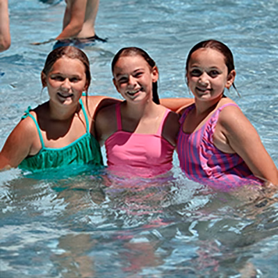 Three smiling girls in colorful swimsuits standing in a pool together, waist-deep in clear water.