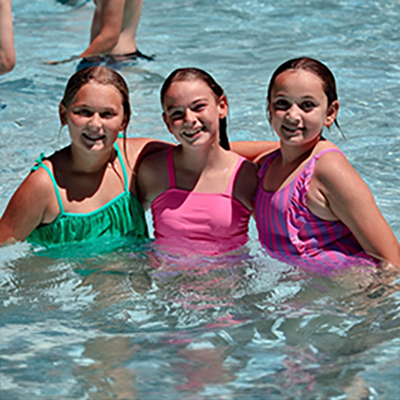 Three smiling girls in colorful swimsuits standing in a pool together, waist-deep in clear water.