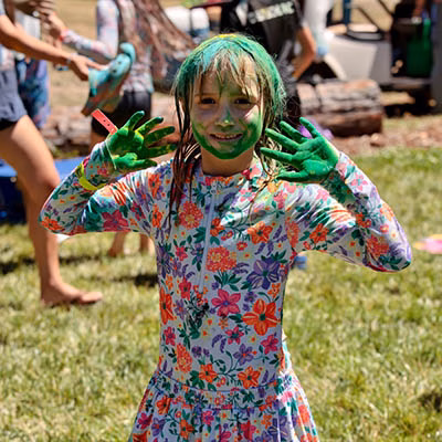 Child with bright green face and hands, wearing a floral dress, posing playfully outdoors during a paint activity.