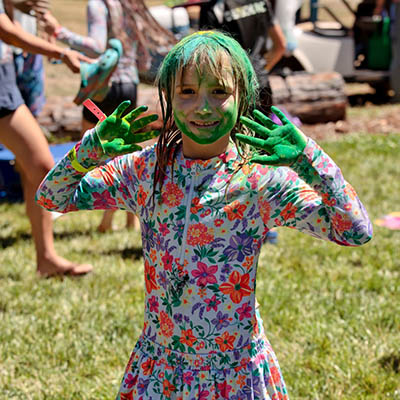 Child with bright green face and hands, wearing a floral dress, posing playfully outdoors during a paint activity.
