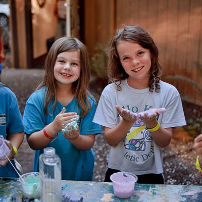 Two smiling girls stand at an outdoor table, playing with colorful slime in cups and bowls.