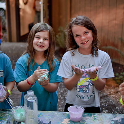 Two smiling girls stand at an outdoor table, playing with colorful slime in cups and bowls.
