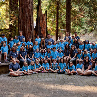 Large group of children and adults posing for a group photo in a forest clearing, many wearing blue shirts.