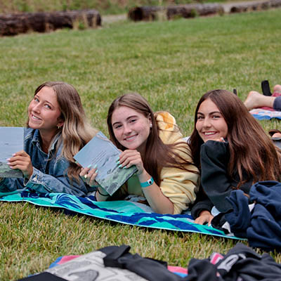 Three young women lie on a blanket on a grassy field, smiling at the camera; one holds a book and the group looks relaxed outdoors.