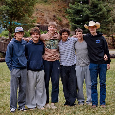 Seven young men standing side by side with arms around each other on a grassy field, wearing casual sweatshirts and pants, with trees and a hillside in the background.