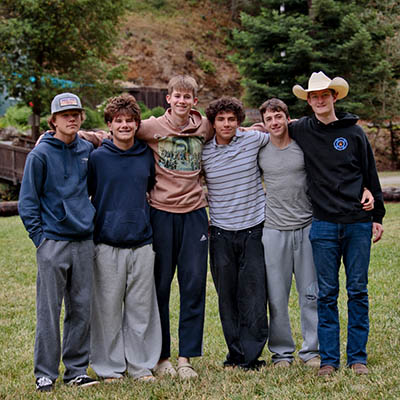 Seven young men standing side by side with arms around each other on a grassy field, wearing casual sweatshirts and pants, with trees and a hillside in the background.