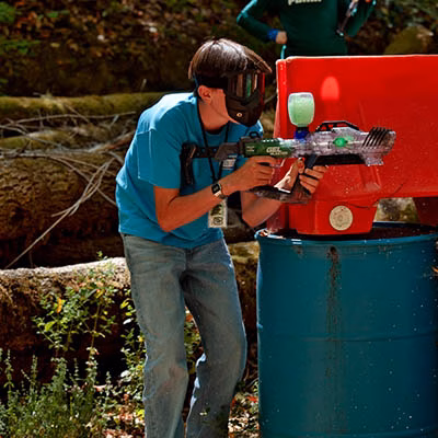 Person in a teal shirt and protective goggles aims a paintball gun at a red barricade during an outdoor game.