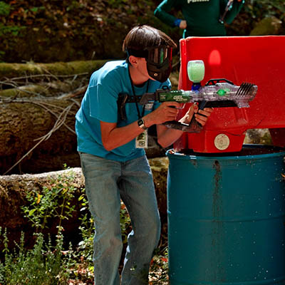 Person in a teal shirt and protective goggles aims a paintball gun at a red barricade during an outdoor game.