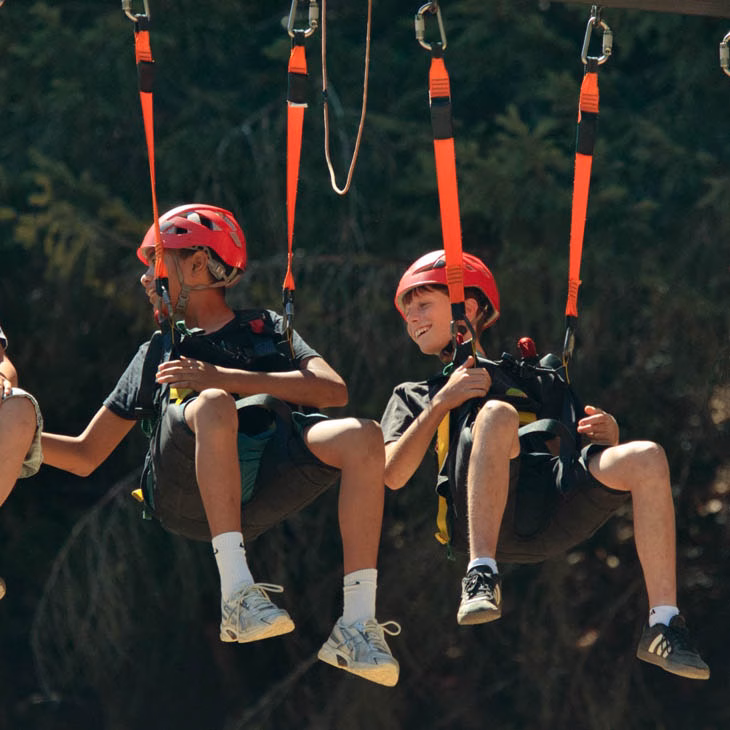 Two children in red helmets sit in harnesses on a zipline ride outdoors, smiling as they glide above the ground with a forest backdrop.