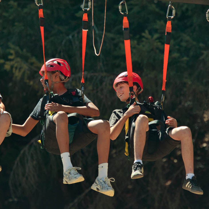 Two children in red helmets sit in harnesses on a zipline ride outdoors, smiling as they glide above the ground with a forest backdrop.