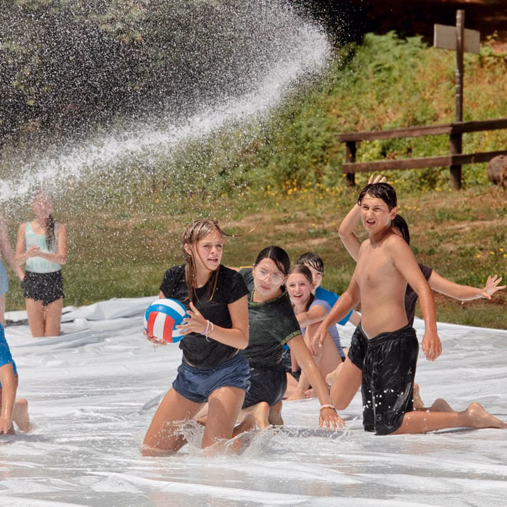 Children playing in a shallow water area, chasing a volleyball as water splashes around them under a spray of water.